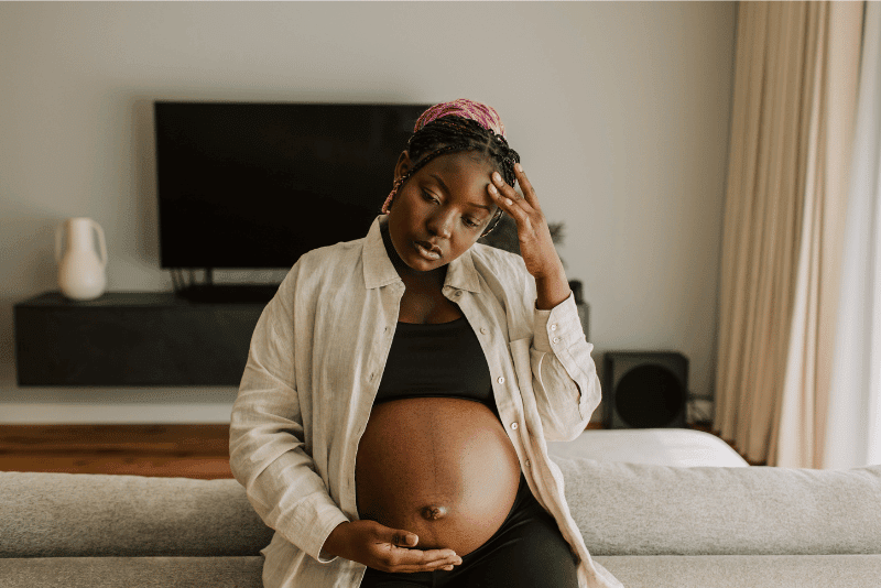Schwangere Frau mit Beschwerden, hält sich den unteren Bauch sanft, im Hintergrund beruhigende Umgebung